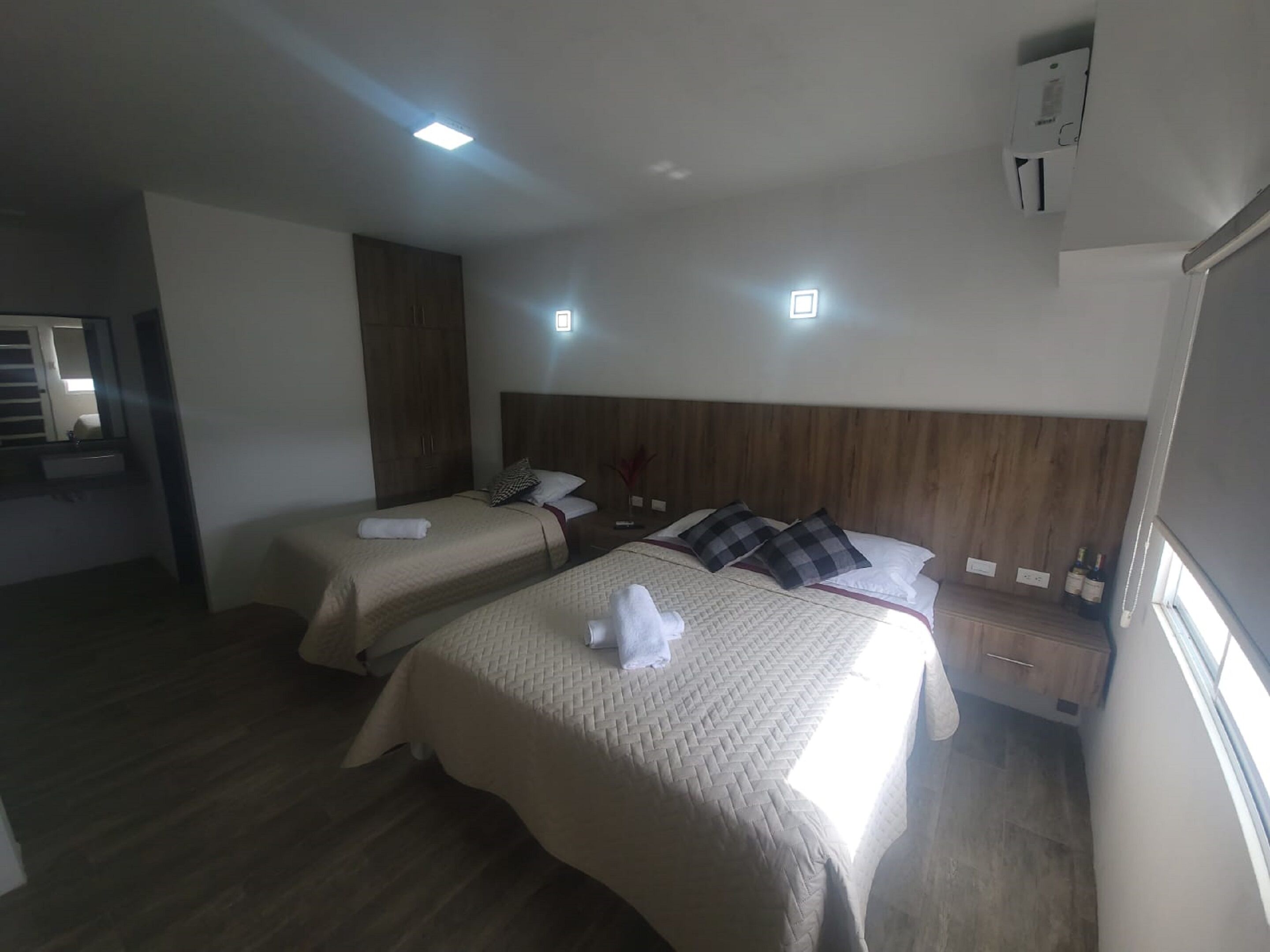 A hotel room with two beds, a wooden headboard, a small bedside table with a lamp, and a window with curtains.