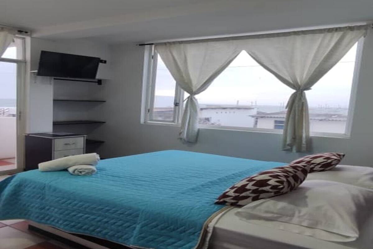 A bedroom with a bed, a TV mounted on the wall, a desk with a remote control, and a window with curtains.
