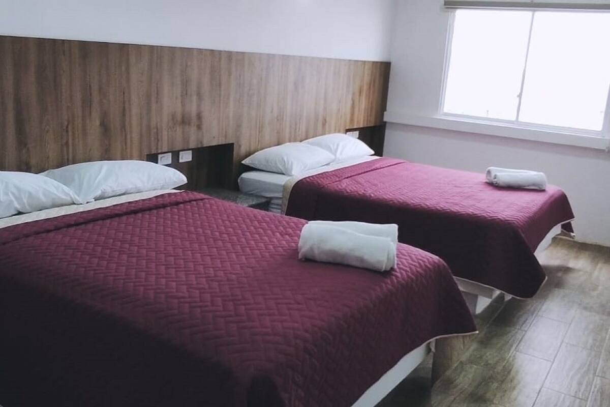 A hotel room with two single beds, each with a red bedspread and white pillows. The room has a wooden headboard and a window with blinds.