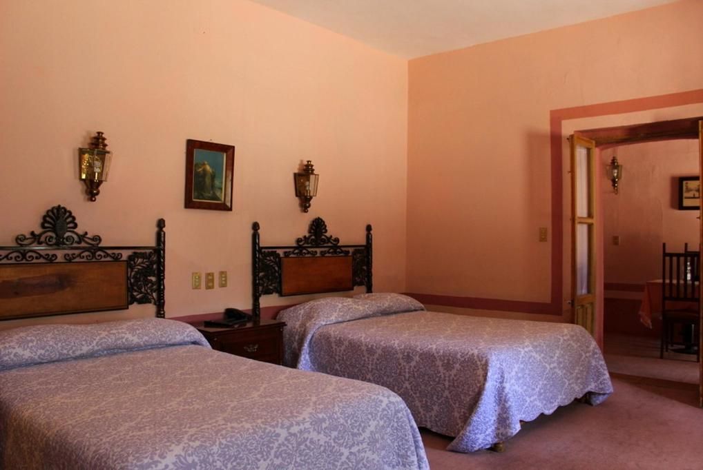 Habitación de hotel con dos camas individuales, cabecero de madera, una mesita de noche y una puerta que da a un comedor.