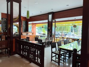 An indoor dining area with tables and chairs, a counter with a menu, and a view of the street outside. - River Grand Hotel Hat Yai (Hat Yai)