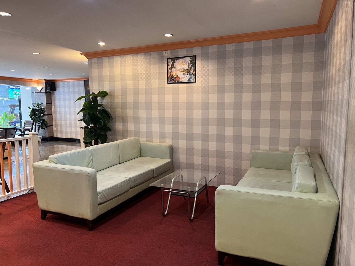 Un vestíbulo de hotel con dos sofás de tonos claros, una mesa de centro de vidrio, una planta grande en maceta y una pared a cuadros.