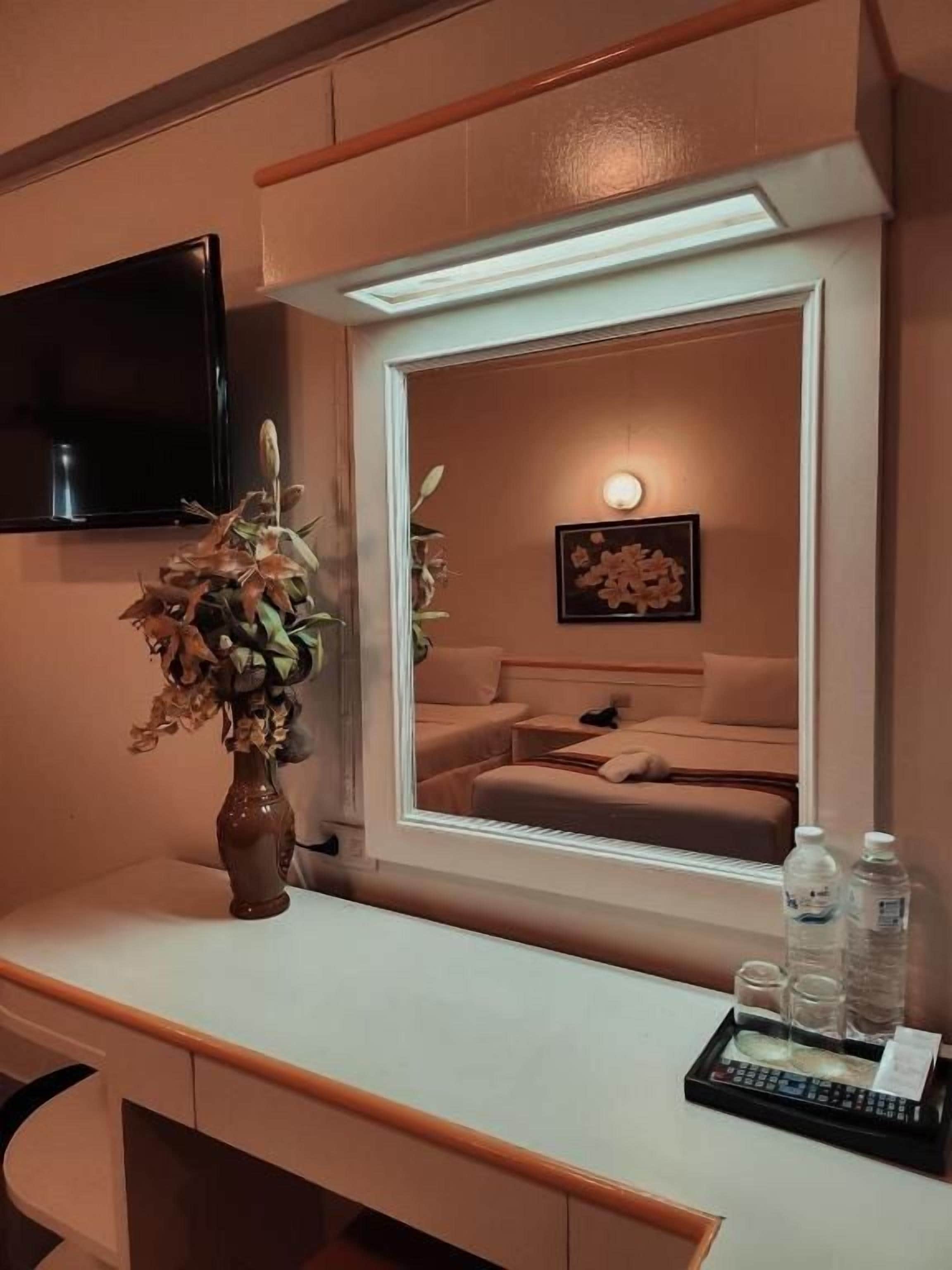 A hotel room with a bed, a bedside table with a vase of flowers, a flat-screen TV mounted on the wall, and a framed artwork above the bed.