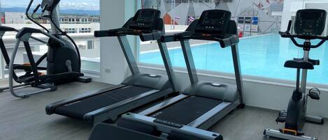A gym with treadmills, an elliptical, and a rowing machine, overlooking a pool and a view of a cityscape.