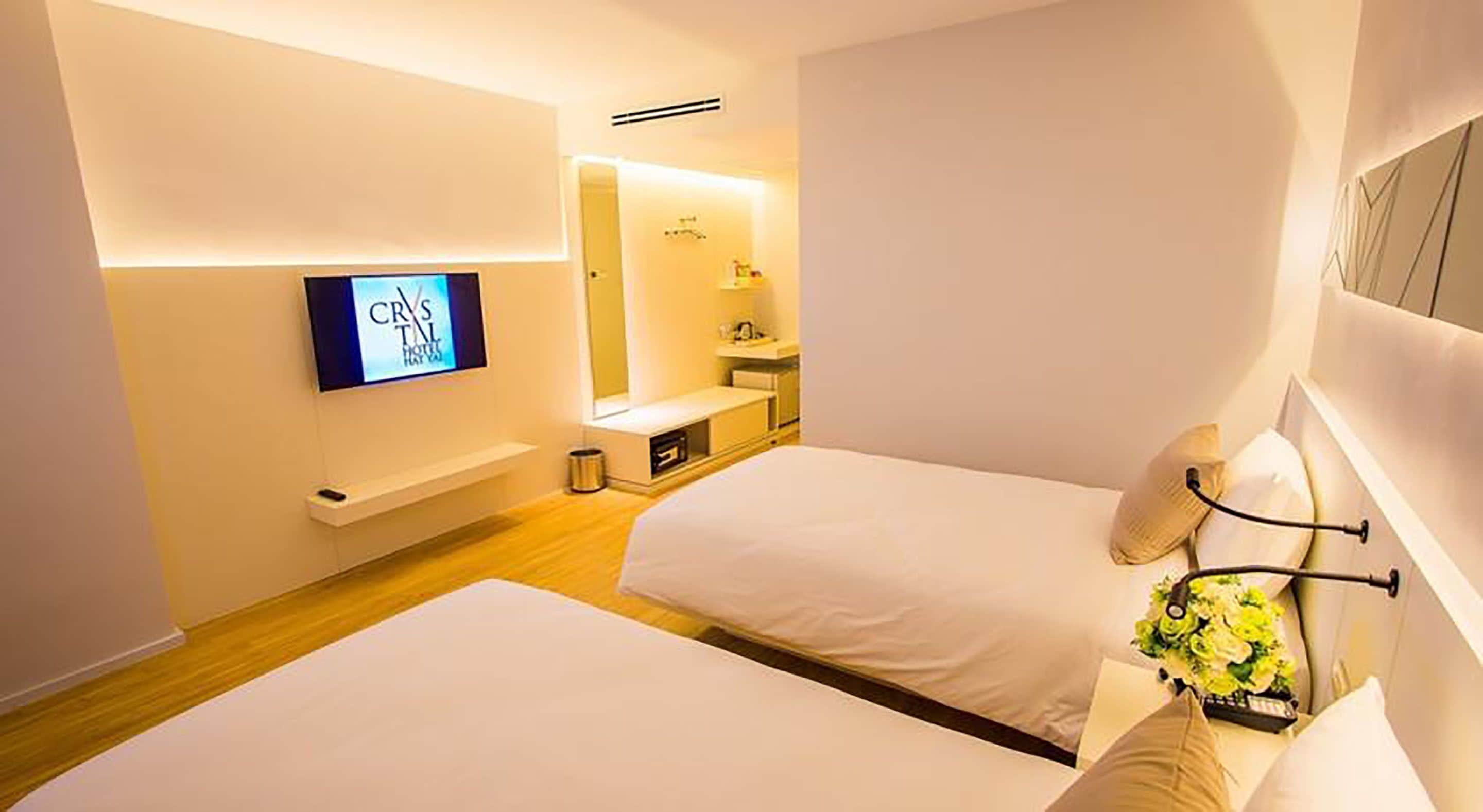 Habitación de hotel con dos camas, un televisor montado en la pared, una mesita de noche con lámpara y una planta pequeña.