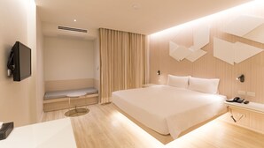Een moderne hotelkamer met een groot bed, een nachtkastje met een telefoon, een flatscreen-tv aan de wand, een zithoek met een bankje en een klein tafeltje, en een groot raam met gordijnen.