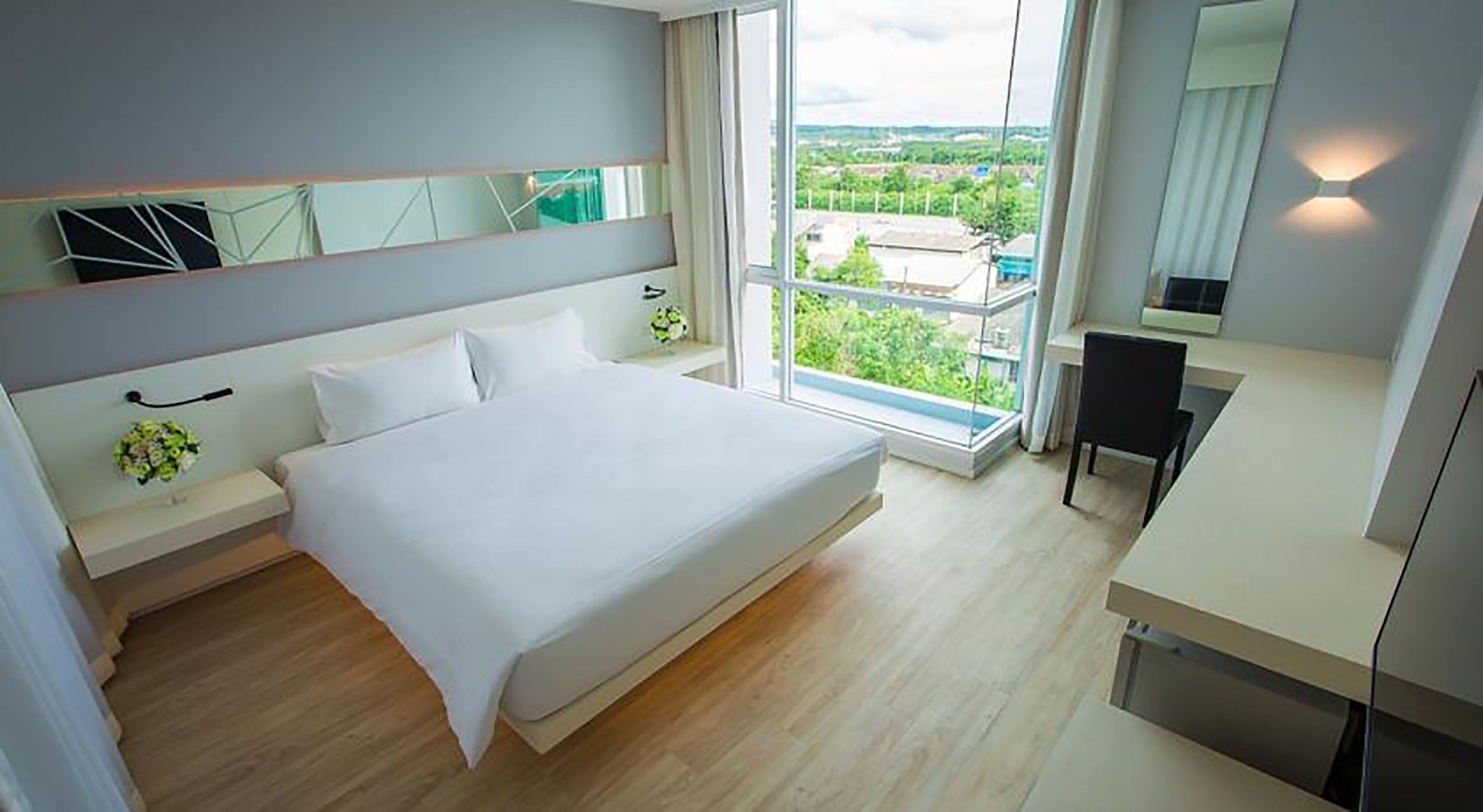 A modern bedroom with a large bed, a desk, and a view of greenery outside.