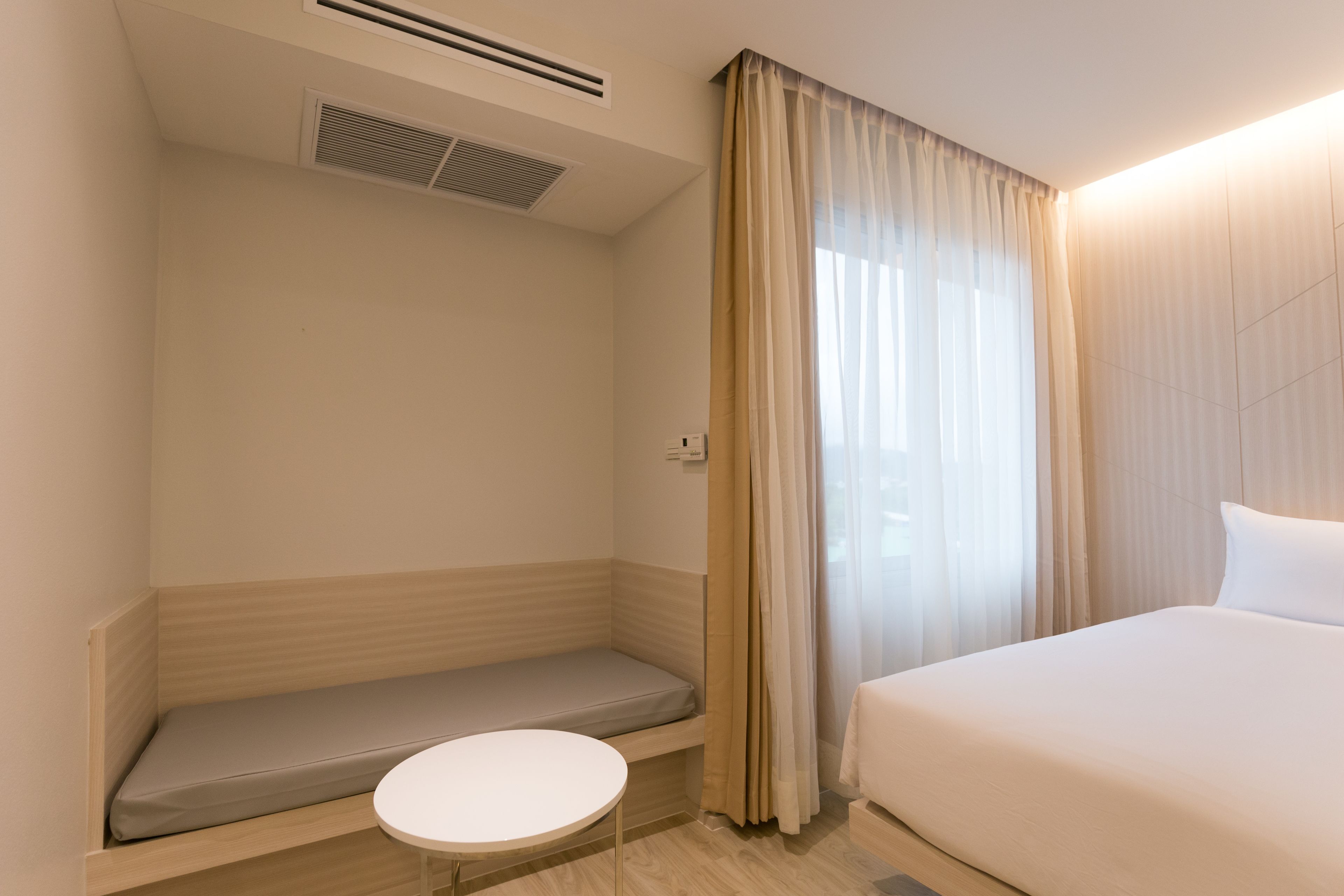 Een hotelkamer met een bed, een klein rond tafeltje, een bankje en een raam met gordijnen.