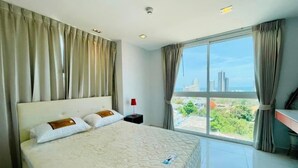A hotel room with a large bed, a nightstand, a red lamp, and a view of the city through large windows. - Art on The Hill by PSR ASIA (Pattaya)