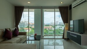 A modern living room with a sofa, a coffee table, a TV, and large windows offering a city view. - Art on The Hill by PSR ASIA (Pattaya)