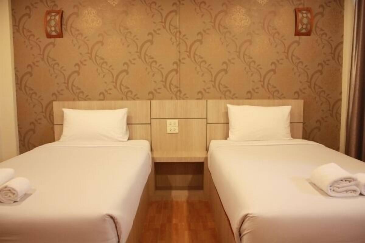 A hotel room with two single beds, wooden headboard, patterned wallpaper, and a nightstand with a lamp.