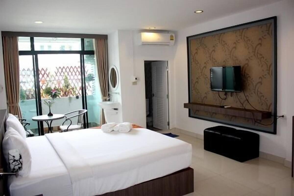 A hotel room with a bed, a TV mounted on the wall, a small table with a chair, and a balcony with a view.