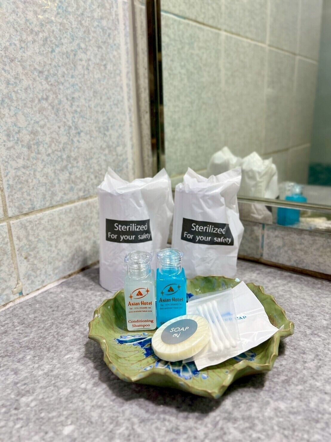A bathroom counter with toiletries including a green leaf-shaped soap dish, a bottle of shampoo, and two bags labeled 'Sterilized for your safety'.