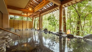 A traditional Japanese onsen with a wooden interior, stone bath, and a view of lush greenery outside.