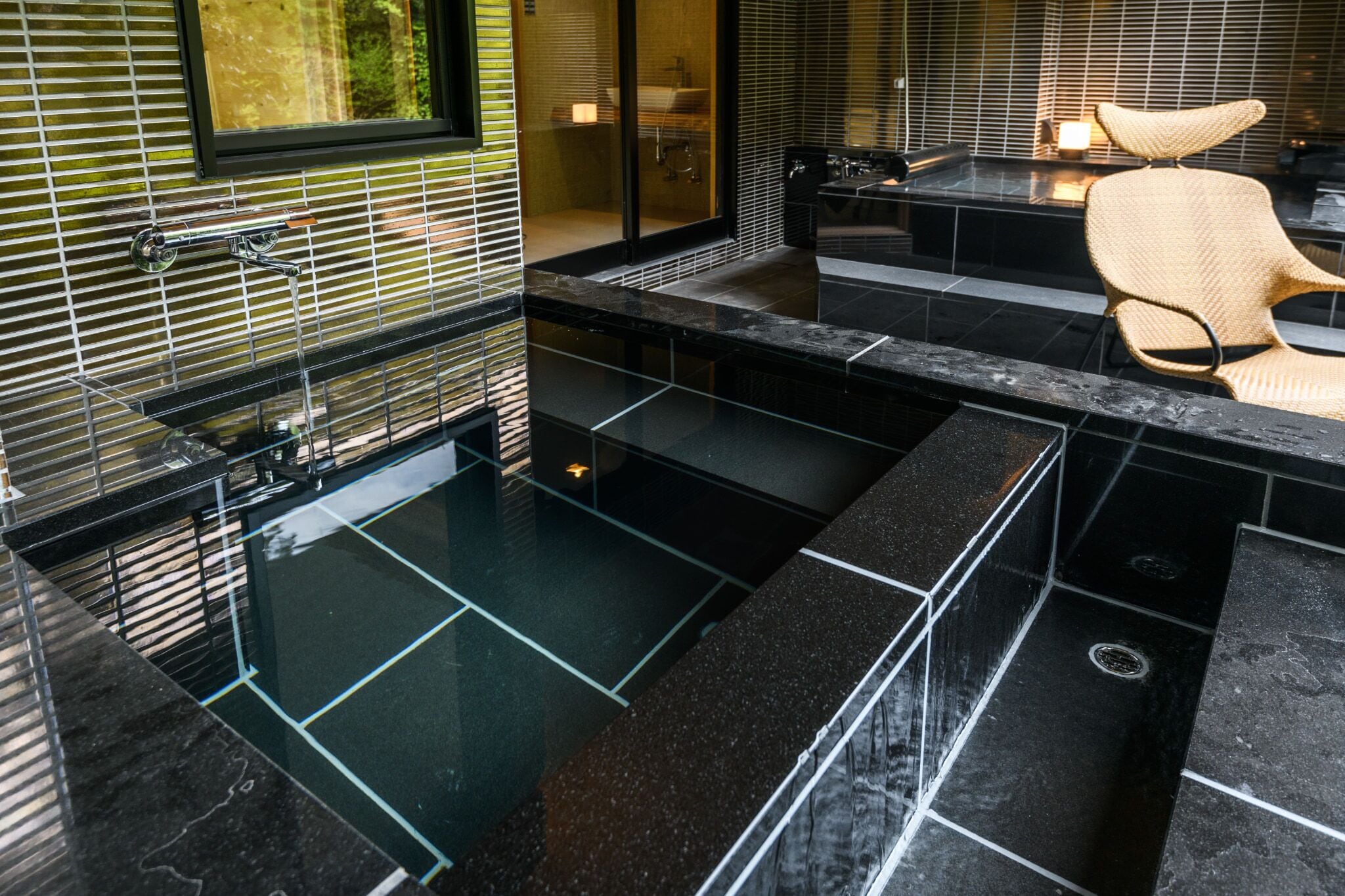 A modern bathroom with a glass-enclosed shower, a black marble countertop, and a woven chair.