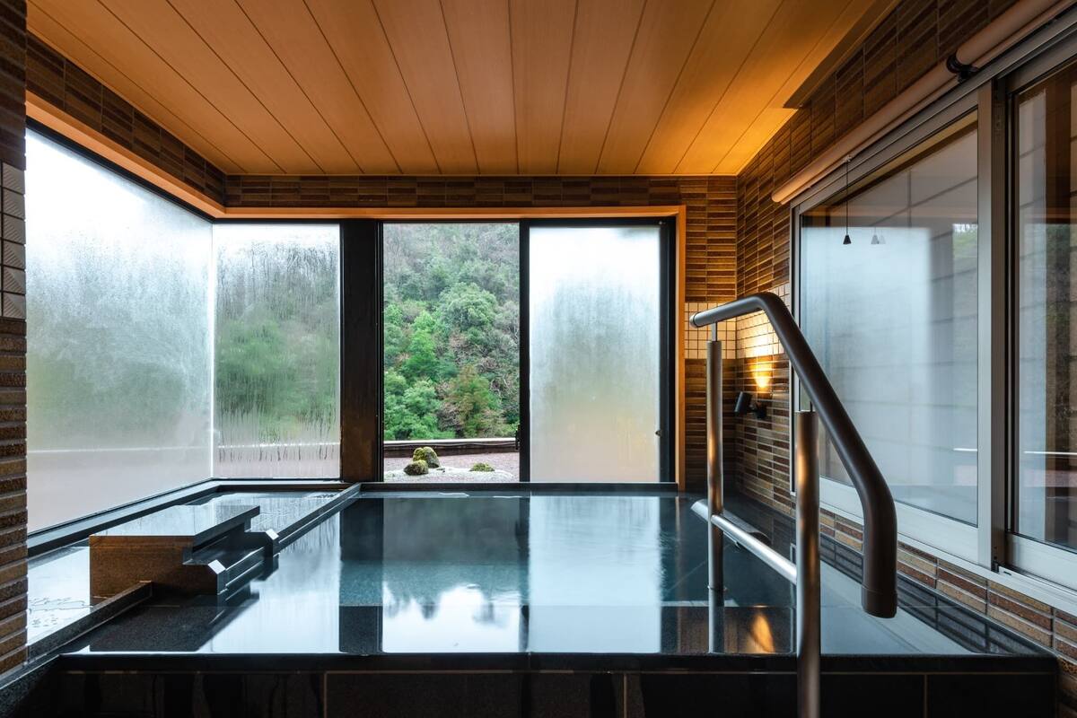 A modern bathroom with a glass-enclosed shower, wooden ceiling, and large windows offering a view of a forested area.