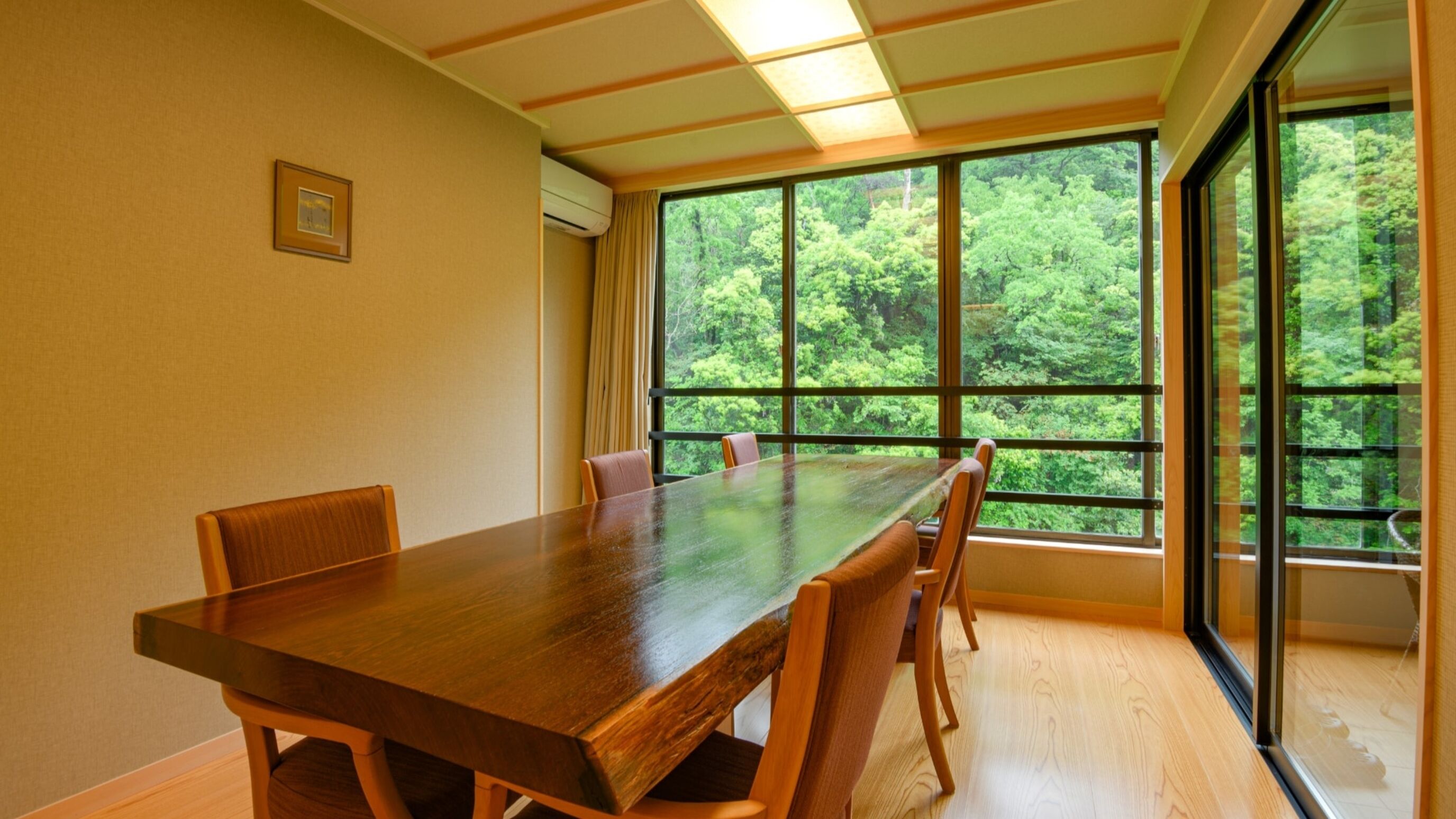 Un salón de conferencias con una larga mesa y sillas, ventanas amplias con vista a un bosque y un cuadro en la pared.