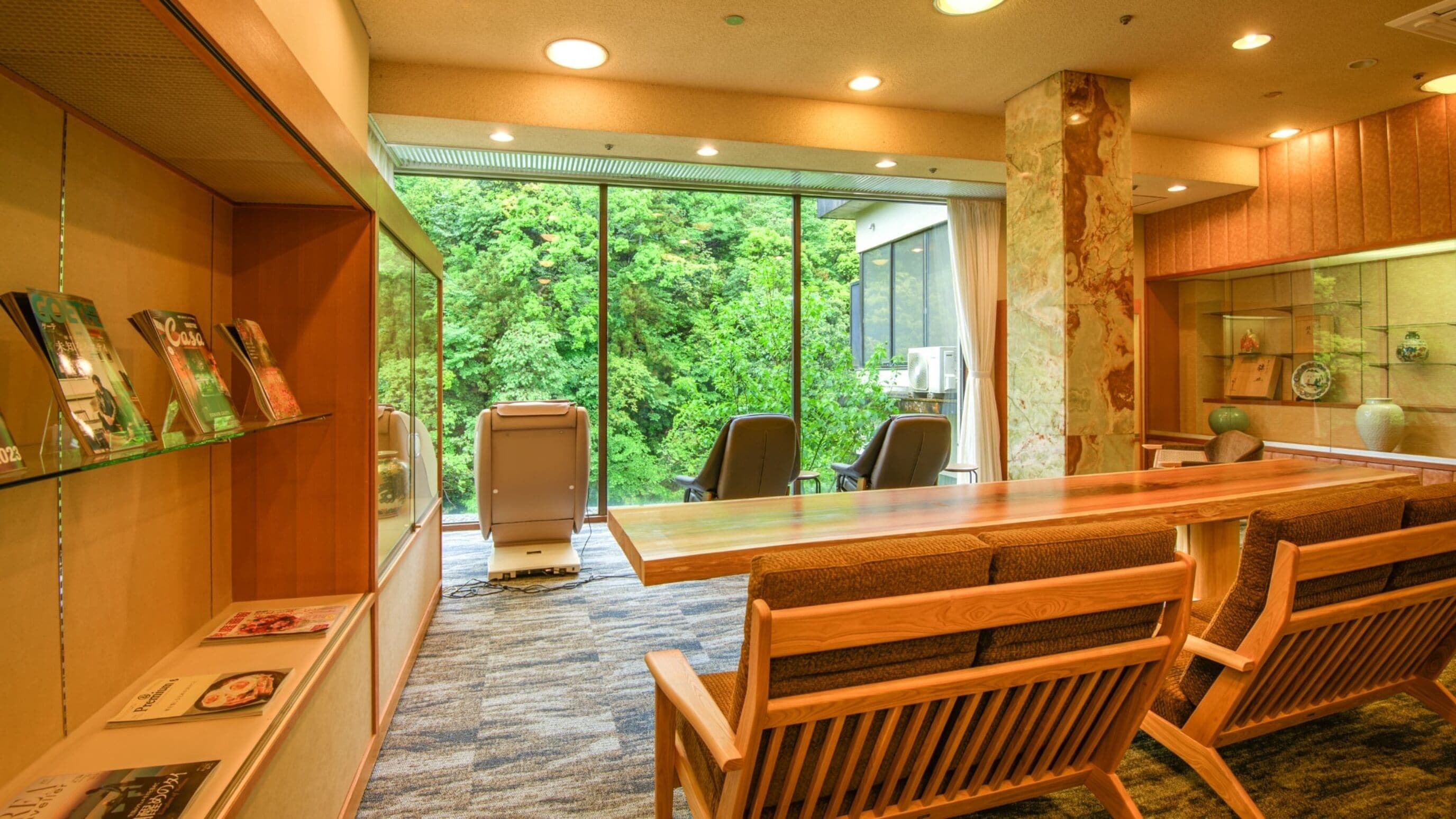 A modern dining area with a long wooden table, several chairs, and a view of lush greenery through large windows.