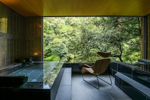 Un interior moderno con una mesa de vidrio, una silla de mimbre y vistas a un bosque a través de amplios ventanales.