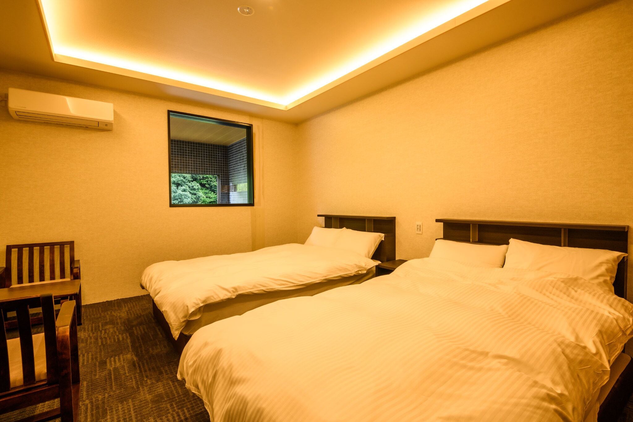 A hotel room with two single beds, a small desk, and a window with a view of greenery.