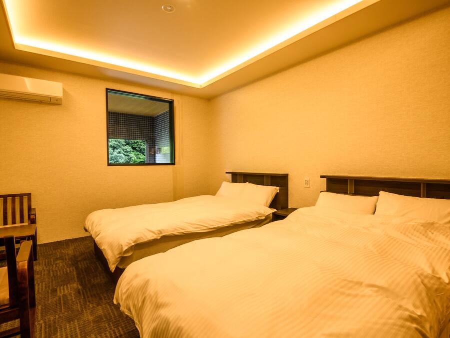 A hotel room with two single beds, a small desk, and a window with a view of greenery.