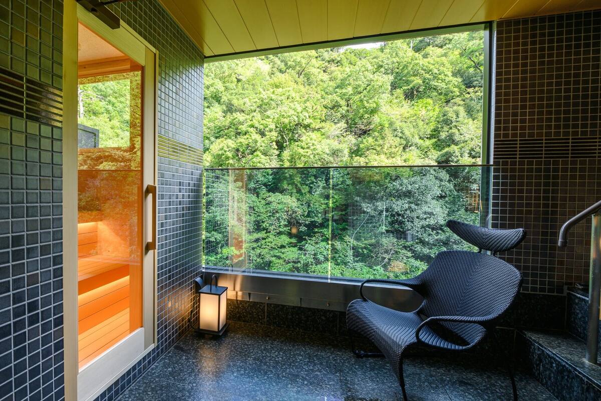 A modern bathroom with a glass door leading to a forest view, a black chair, and tiled walls.