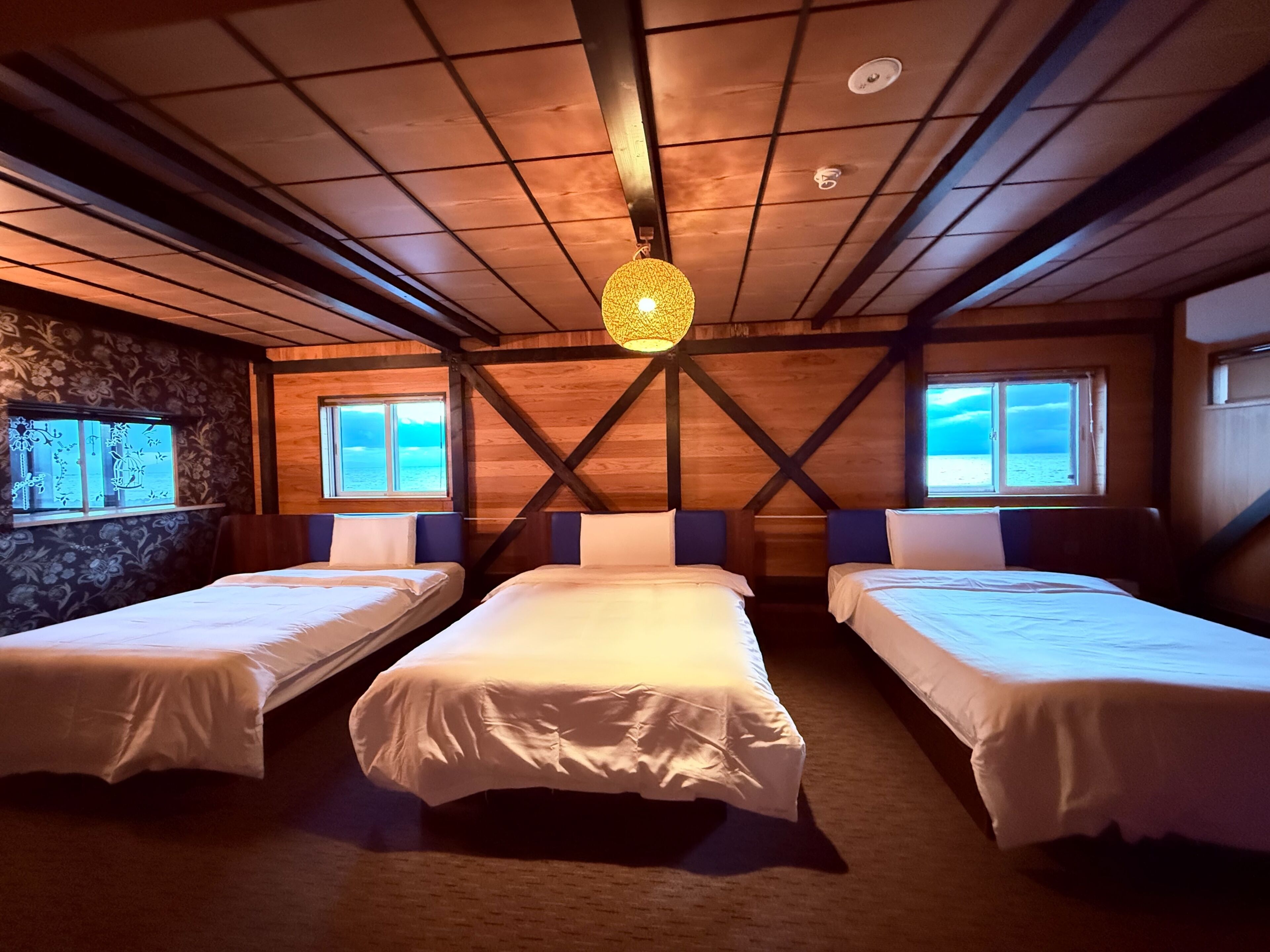 A cabin-style room with three single beds, wooden walls, a stone accent wall, and large windows with a view of the sea.