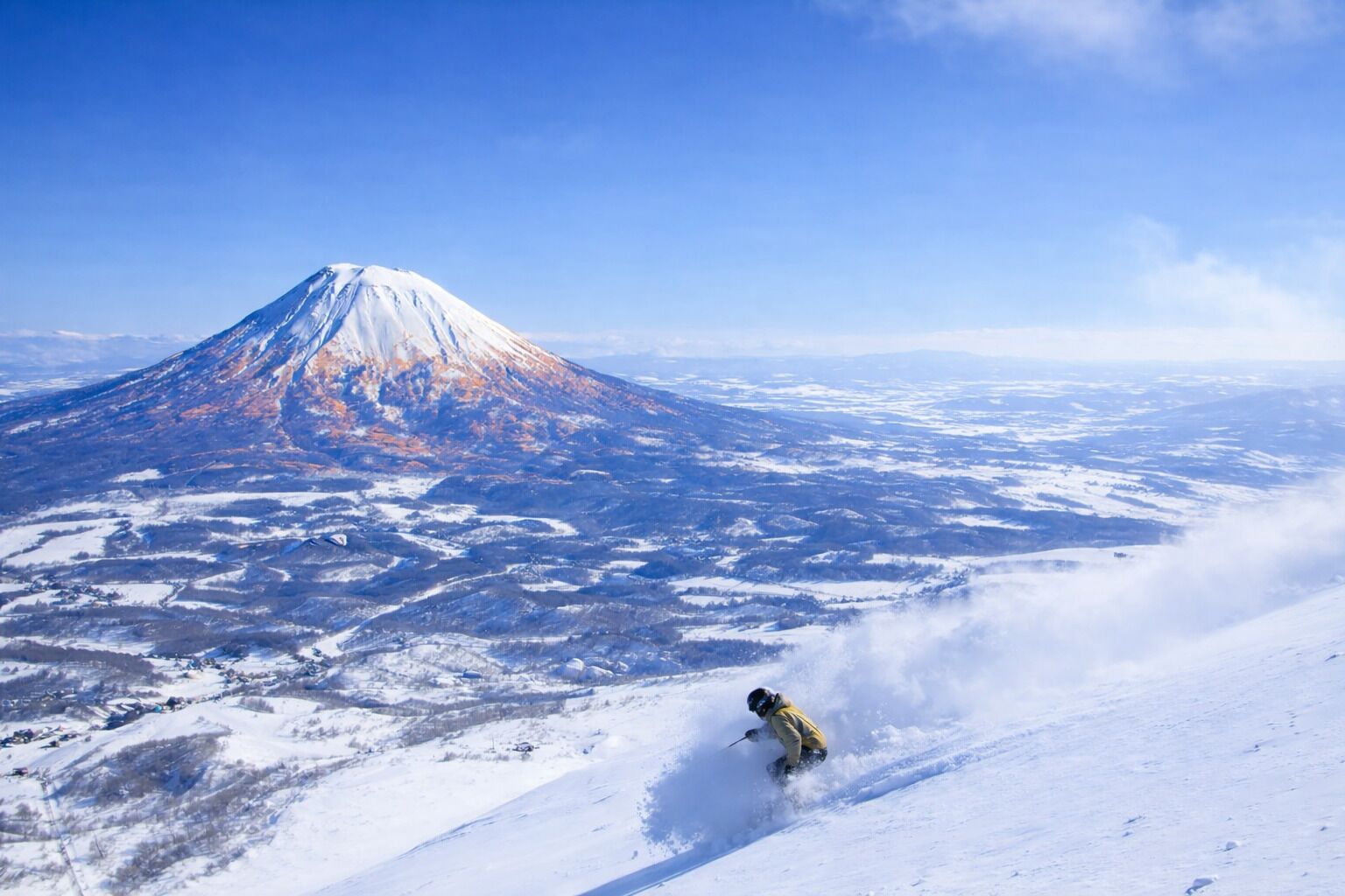 A snow-covered landscape with a prominent mountain in the background and a skier in the foreground.