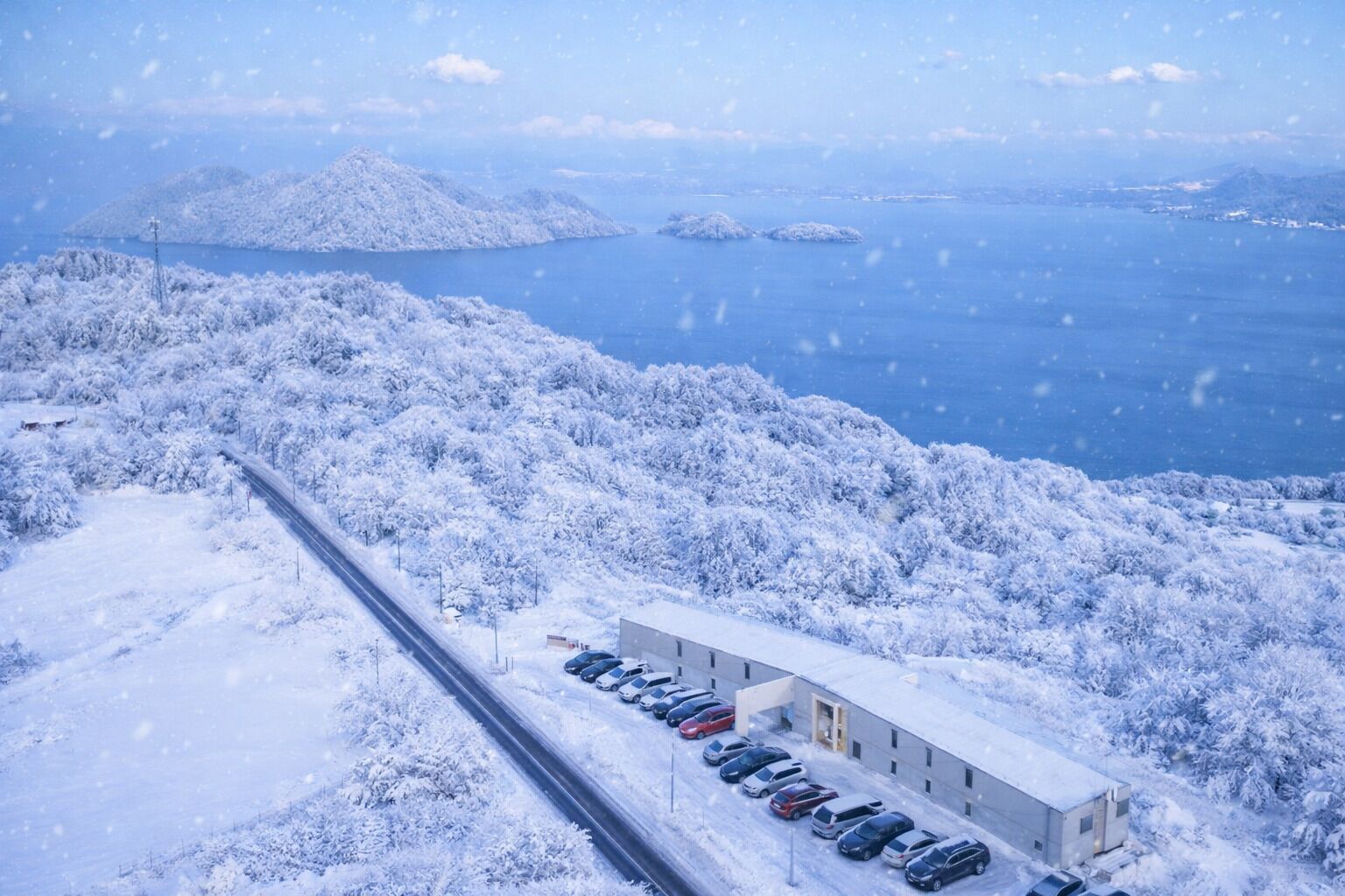 A snow-covered landscape with a road, a building with a parking lot, and a body of water in the background.