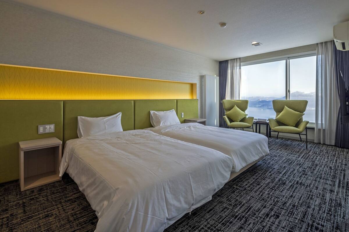 A hotel room with two beds, a view of the sea, a green headboard, and a small table with a chair.