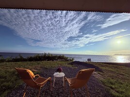 Una terraza con dos sillas de mimbre y una mesita, con vistas a la costa, el océano y un cielo despejado.