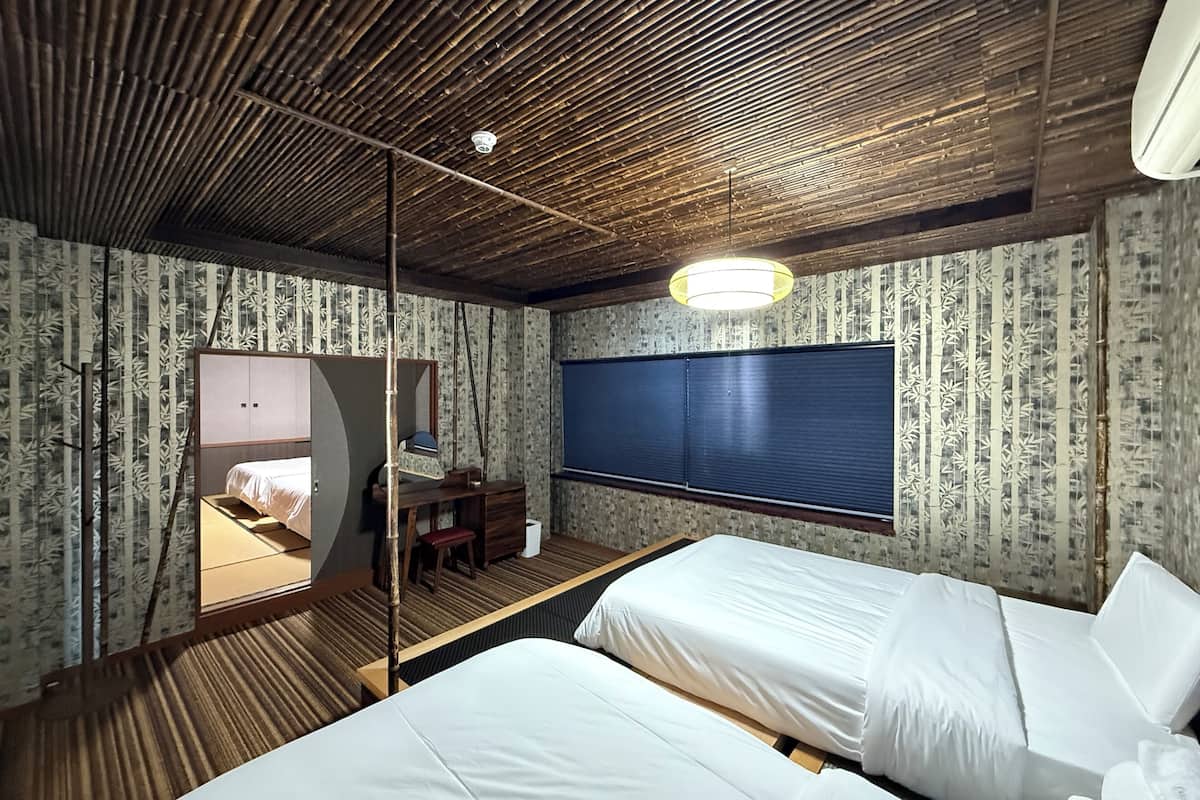 A room with two beds, a desk, a mirror, and a window with blinds.