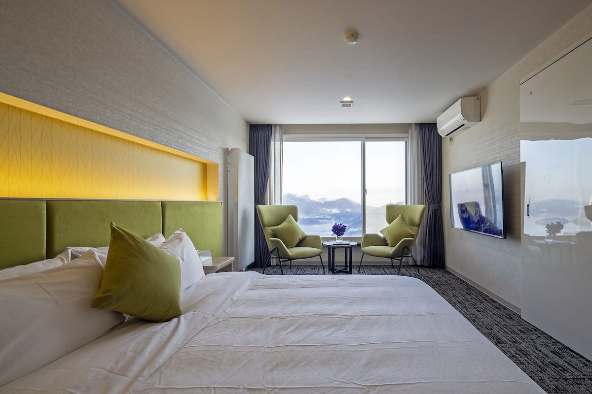 A hotel room with a large bed, two armchairs, a small table, a TV, and a view of mountains through the window.