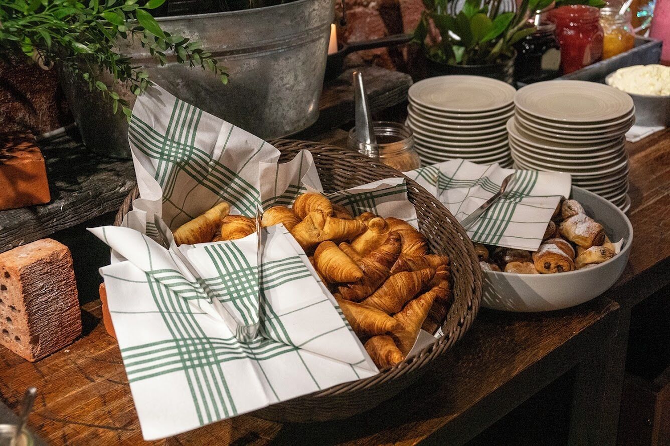 Un buffet de petit-déjeuner avec des croissants et du pain, des piles d’assiettes et un seau en métal.