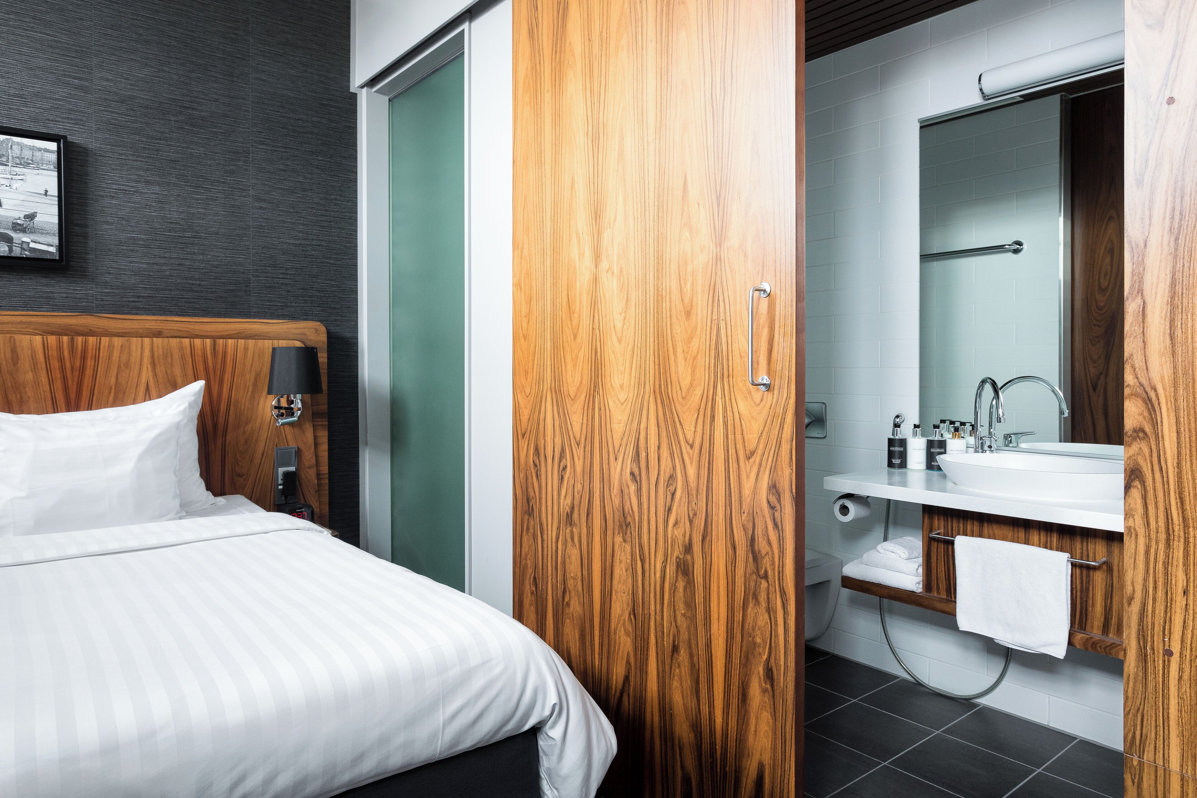 A hotel room with a wooden headboard, a bed with white linens, a wooden wardrobe, a bathroom with a sink, mirror, and towel rack.