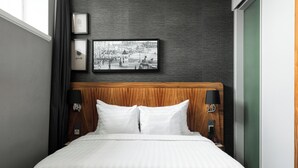 Un lit bien fait, avec du linge de lit blanc, un mur texturé de couleur sombre et, au-dessus, une photographie en noir et blanc encadrée.