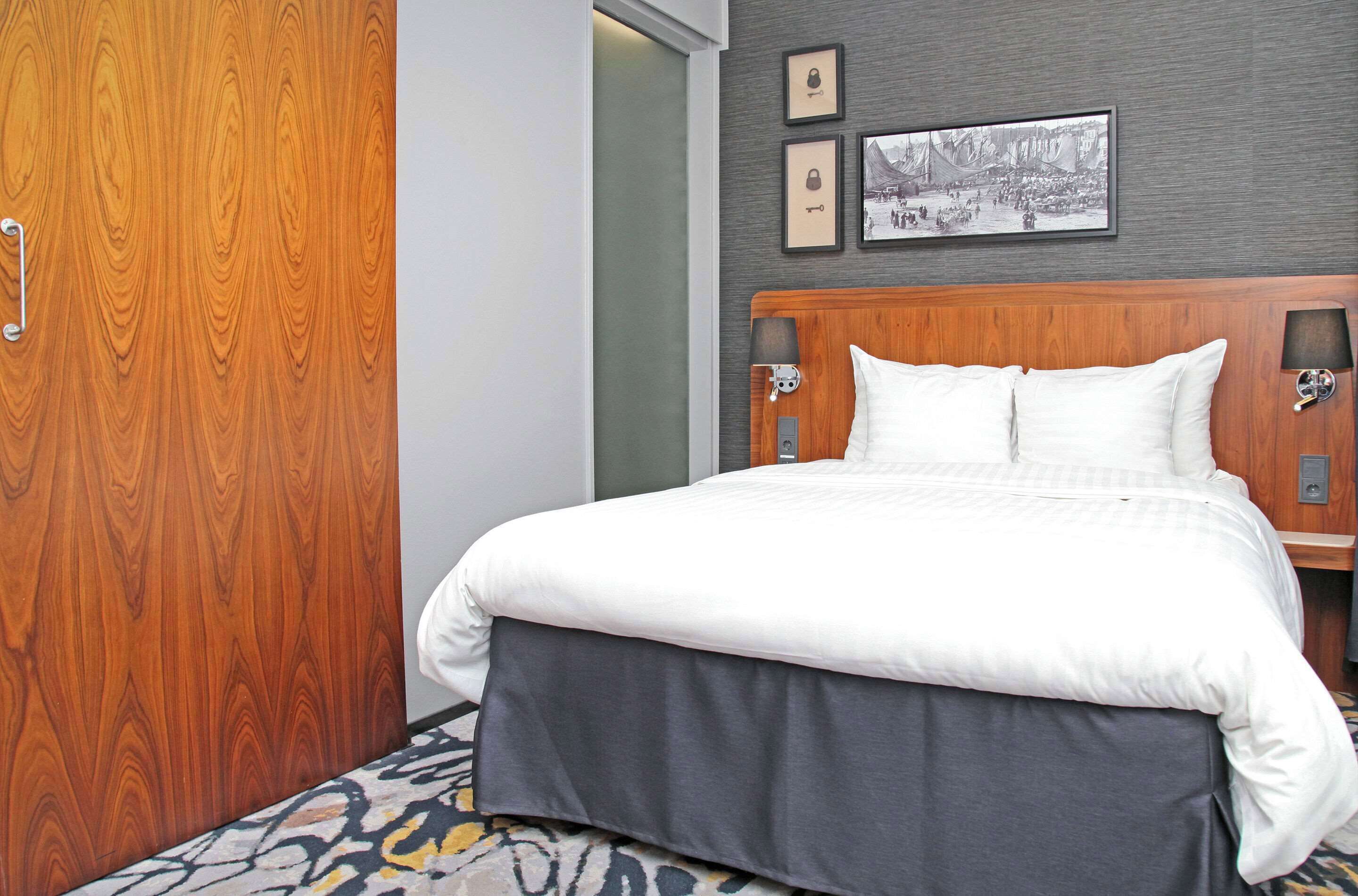Hotelkamer met een houten hoofdeinde, een groot bed met witte beddengoed, een bed met grijze onderrok en een kamerbreed tapijt. Er hangen ingelijste foto’s aan de muur en een deur met een handgreep.