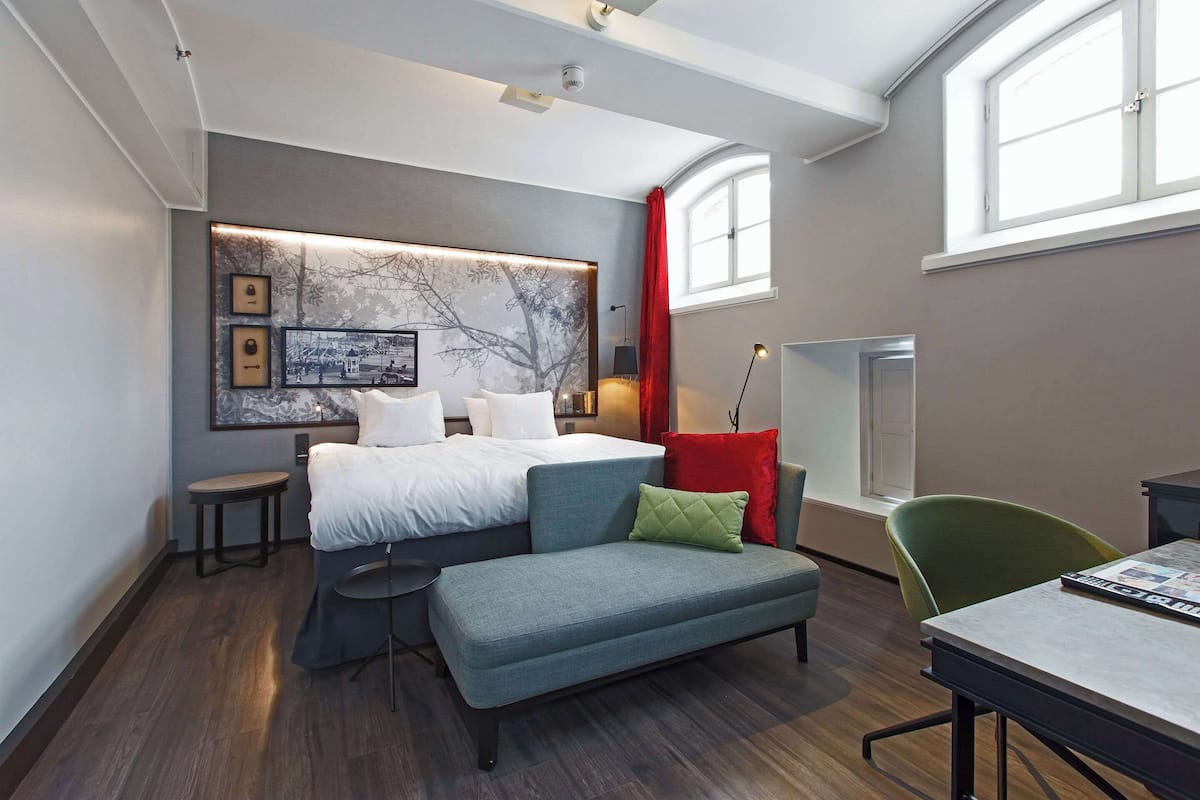 A modern bedroom with a large bed, a sofa, a desk, and a chair. There is a window with red curtains and a view of the outside.