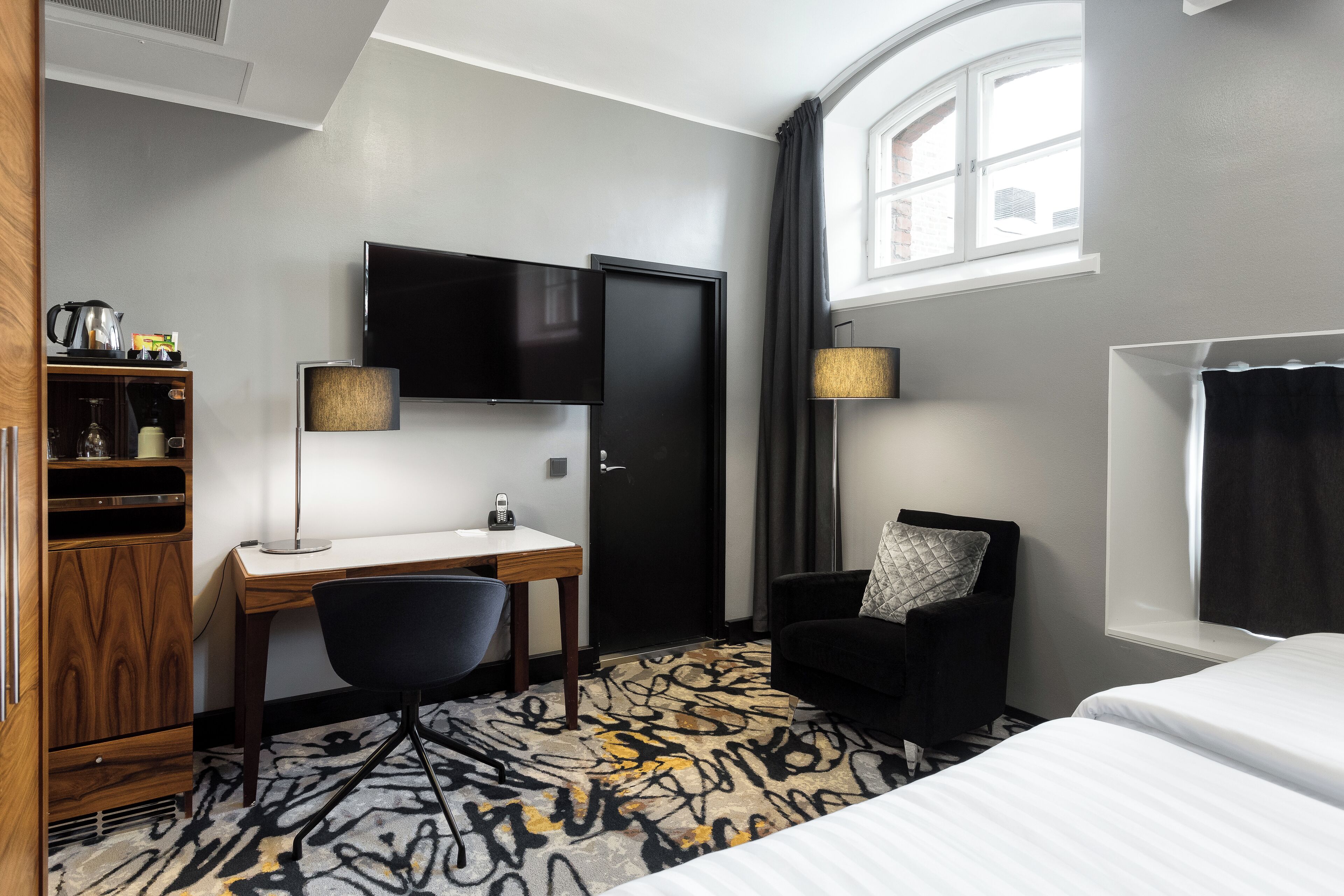 Ein Hotelzimmer mit Schreibtisch, Stuhl, Bett, Fernseher und einem Fenster mit Vorhängen.