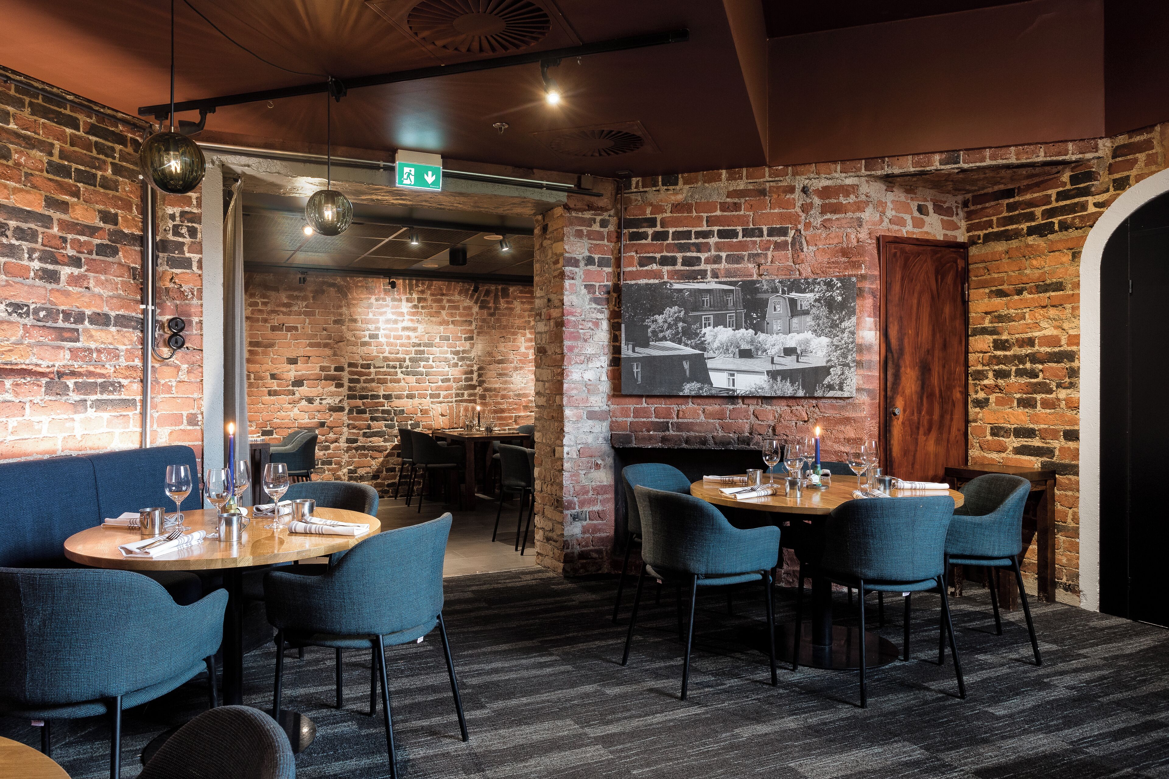 A restaurant with brick walls, blue upholstered chairs, and a dining table set with wine glasses and cutlery.