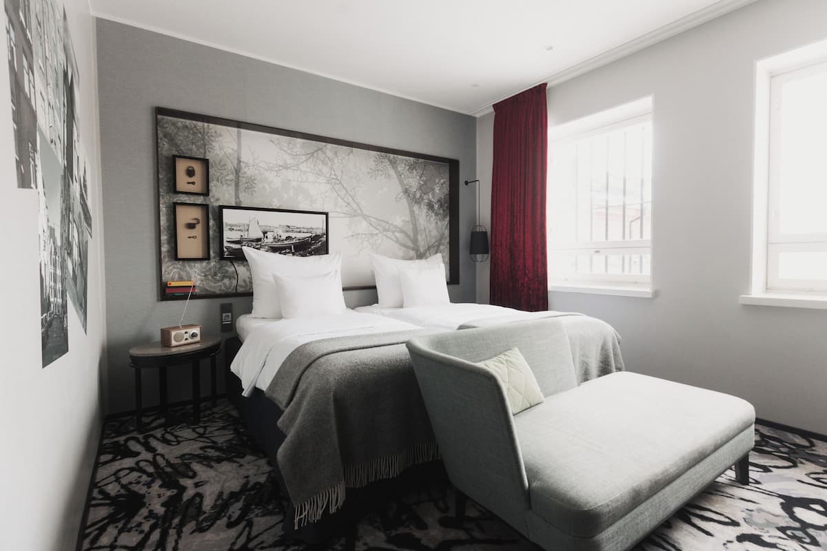 A modern hotel room with a large bed, a side table, a radio, and a view of the outside through a window.