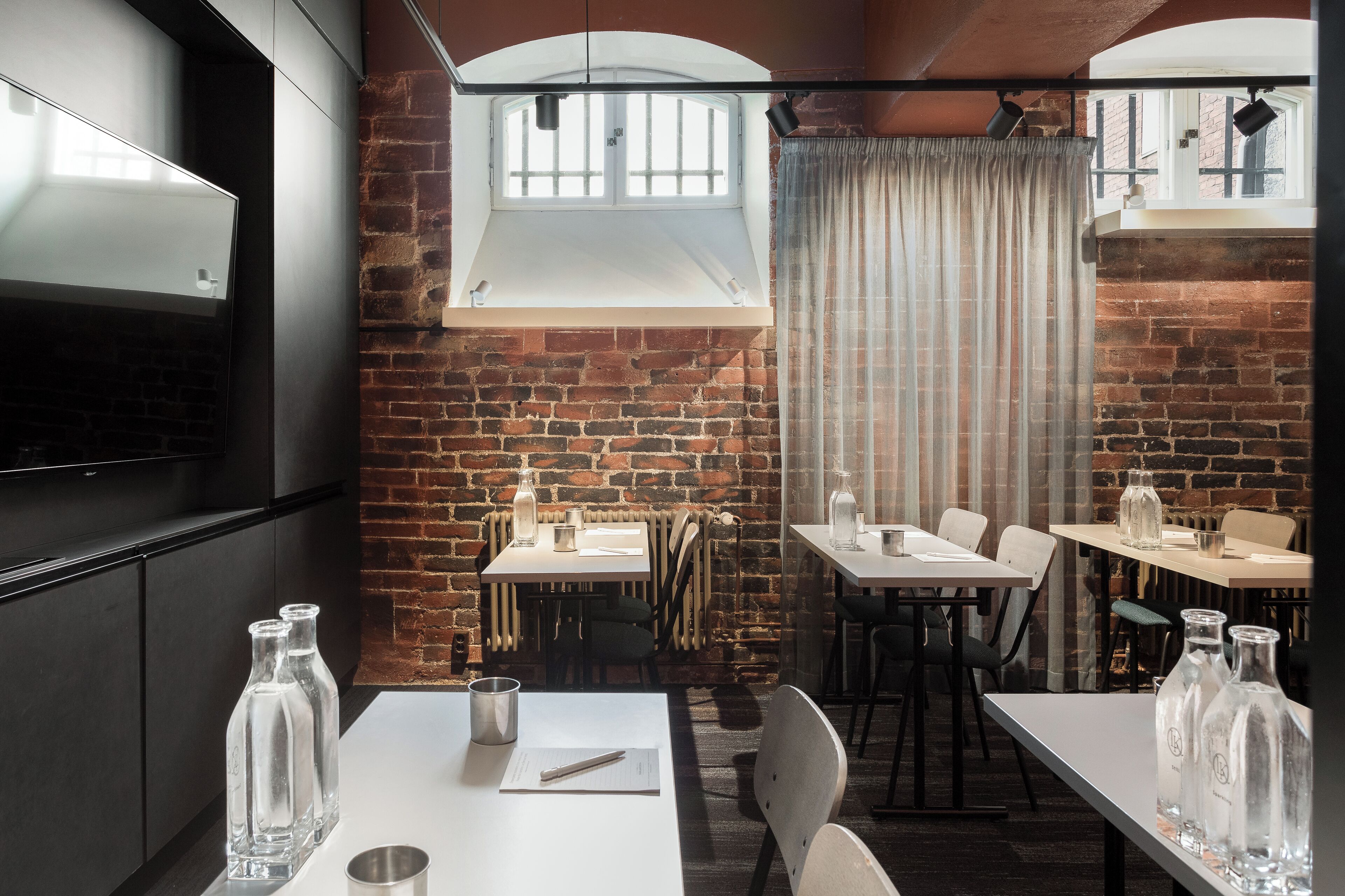 Un restaurante con paredes de ladrillo, mesas y sillas blancas. Hay botellas sobre las mesas y un televisor en la pared.