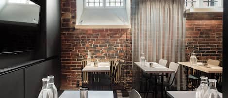 Un restaurante con paredes de ladrillo, mesas y sillas blancas. Hay botellas sobre las mesas y un televisor en la pared.