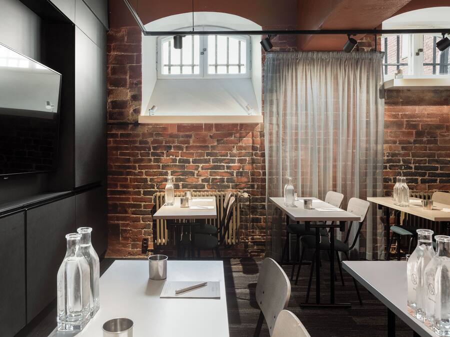 Un restaurante con paredes de ladrillo, mesas y sillas blancas. Hay botellas sobre las mesas y un televisor en la pared.