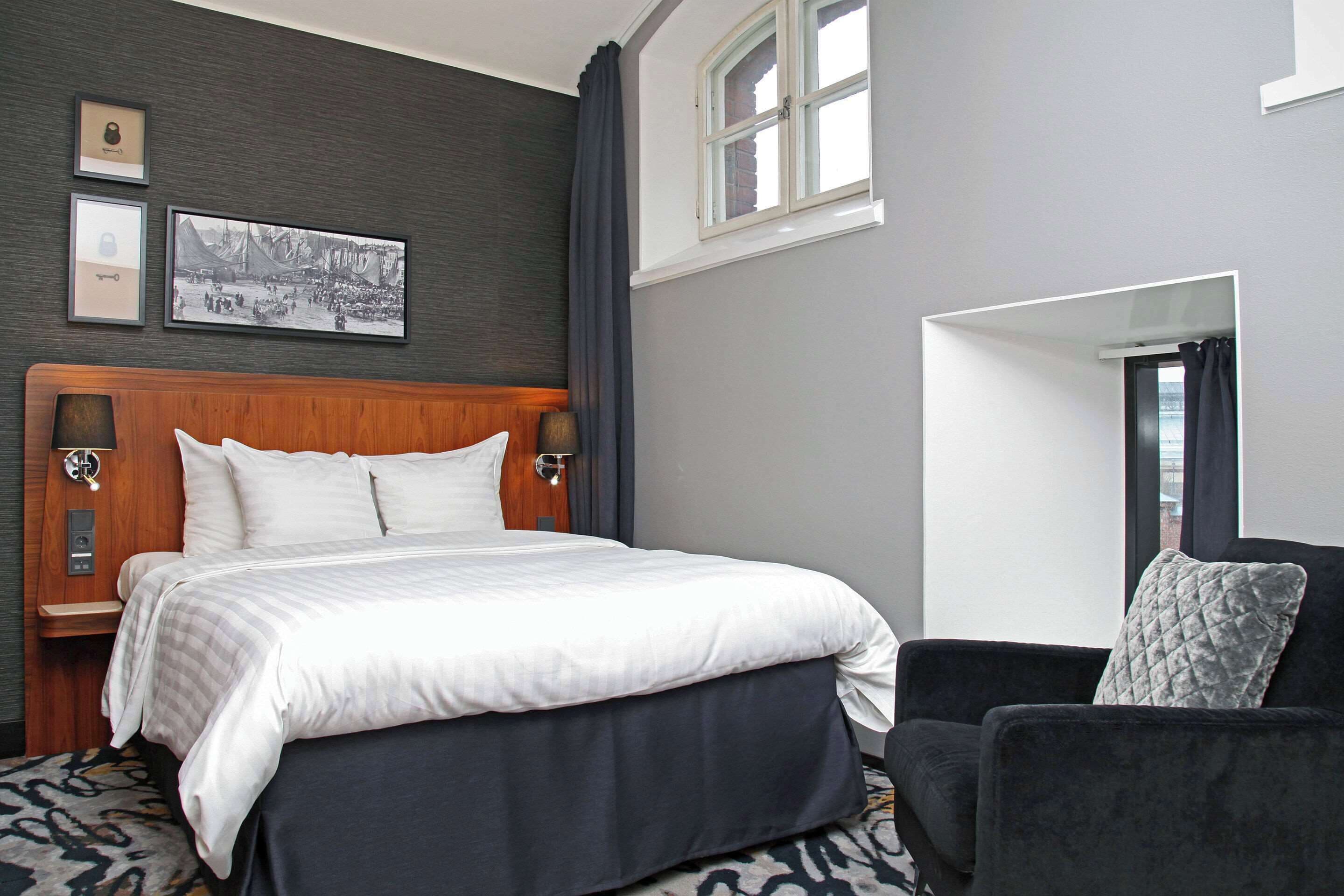 Habitación de hotel con una cama grande, cabecera de madera, un sillón negro y una ventana con cortinas.