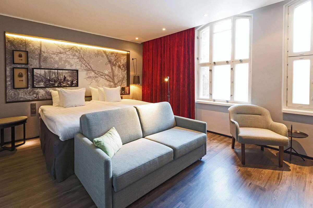 A modern hotel room with a bed, a sofa, a chair, and a large window.
