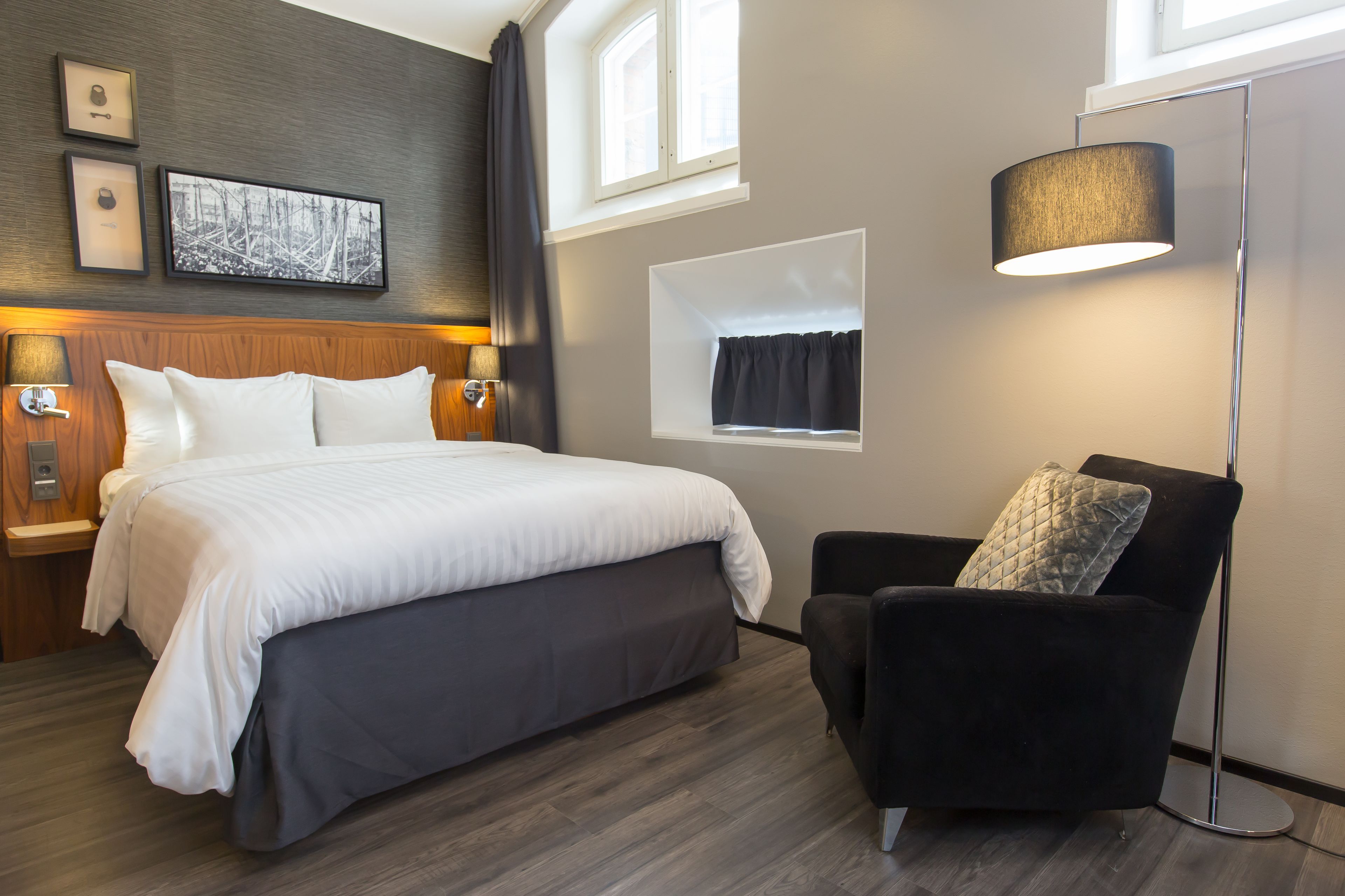 Une chambre d’hôtel moderne avec un grand lit, un fauteuil noir, une lampe sur pied et une fenêtre avec des rideaux.