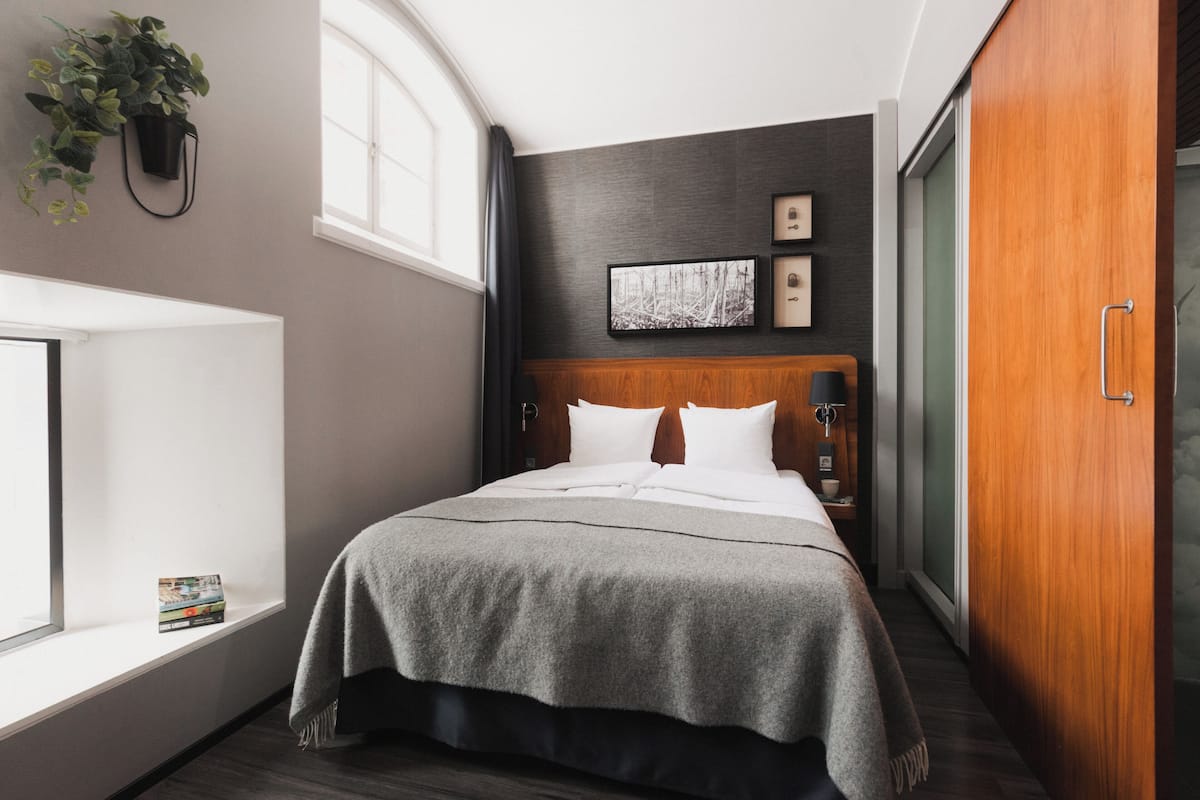 A hotel room with a bed, a window, a plant, and a wooden wardrobe.