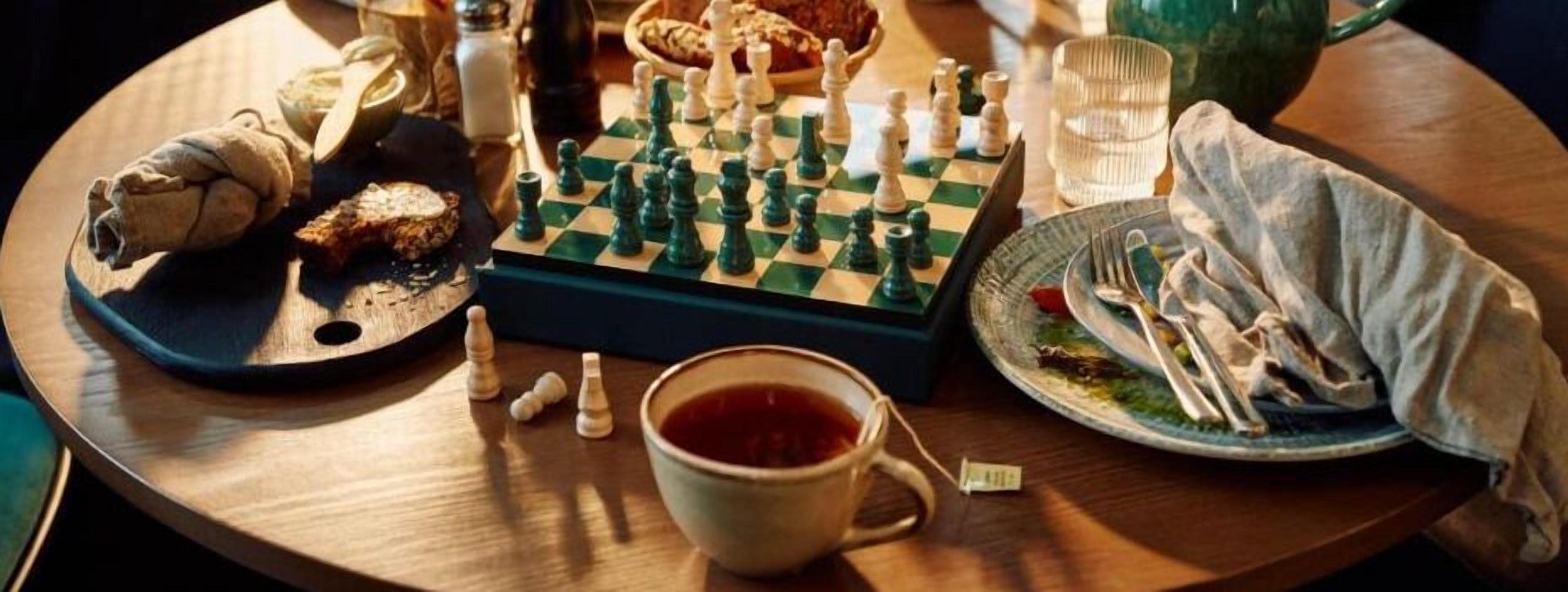 A round wooden table with a chessboard, a cup of tea, a plate with food, and a salt shaker.