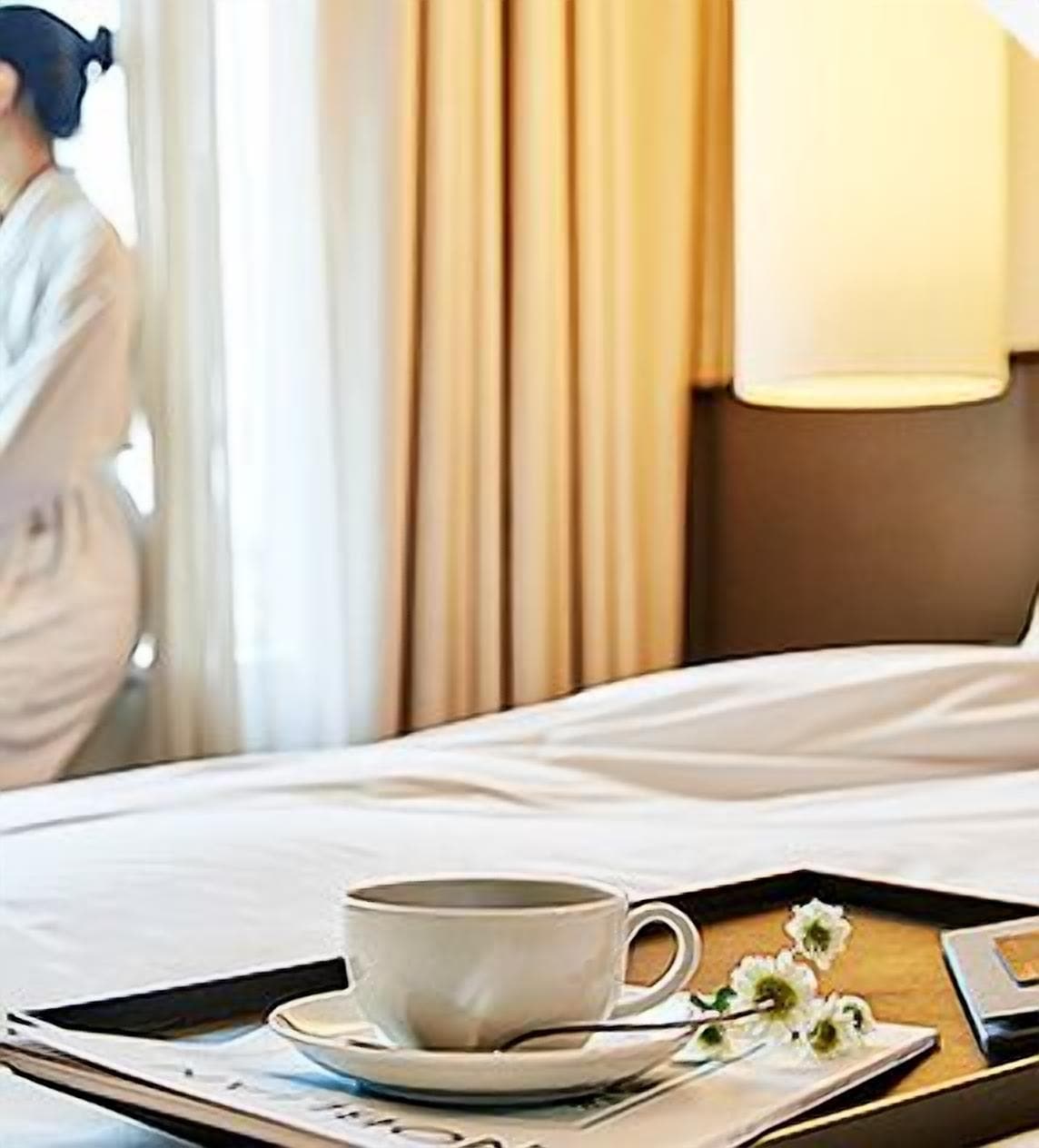Una habitación de hotel con cama, una taza de café en un atril y una lámpara en la mesita de noche.